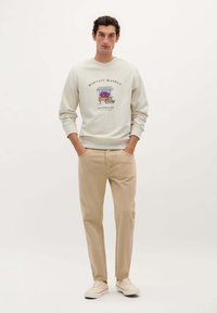Sudadera gris claro con un gráfico de un carrito de mercado y "HARVEST MARKET SATURDAYS". Chinos beige y zapatillas crema, con las manos en los bolsillos.
