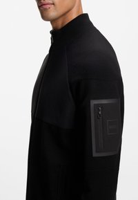 Μαύρο μπουφάν με φερμουάρ και ψηλό γιακά, με υφή στο πάνω μέρος. Διαθέτει τσέπη με φερμουάρ στο αριστερό μανίκι με λαστιχένιο λογότυπο.