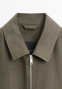 Olivengrøn skjortekrave med synlig syning og en sølvfarvet metal lynlås, delvist lynet op foran i midten.