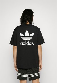 Musta puuvillapaita, jossa on suuri valkoinen Adidas-logo ja kolme valkoista raitaa sen yläpuolella, yhdistettynä kuvioituihin mustiin shortseihin.