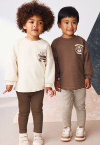 To børn står sammen iført afslappet tøj. Den ene har en cremefarvet sweatshirt med en brun grafik og brune leggings, mens den anden har en mørkebrun sweatshirt og stribede leggings. Begge bærer lysefarvede sneakers.