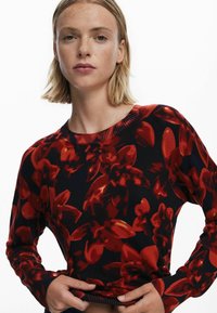 Hosszú ujjú pulóver, amely sötét háttérrel és nagy piros virágmintákkal rendelkezik. Az anyaga kötöttnek tűnik, bordás nyakkivágással és mandzsettákkal.