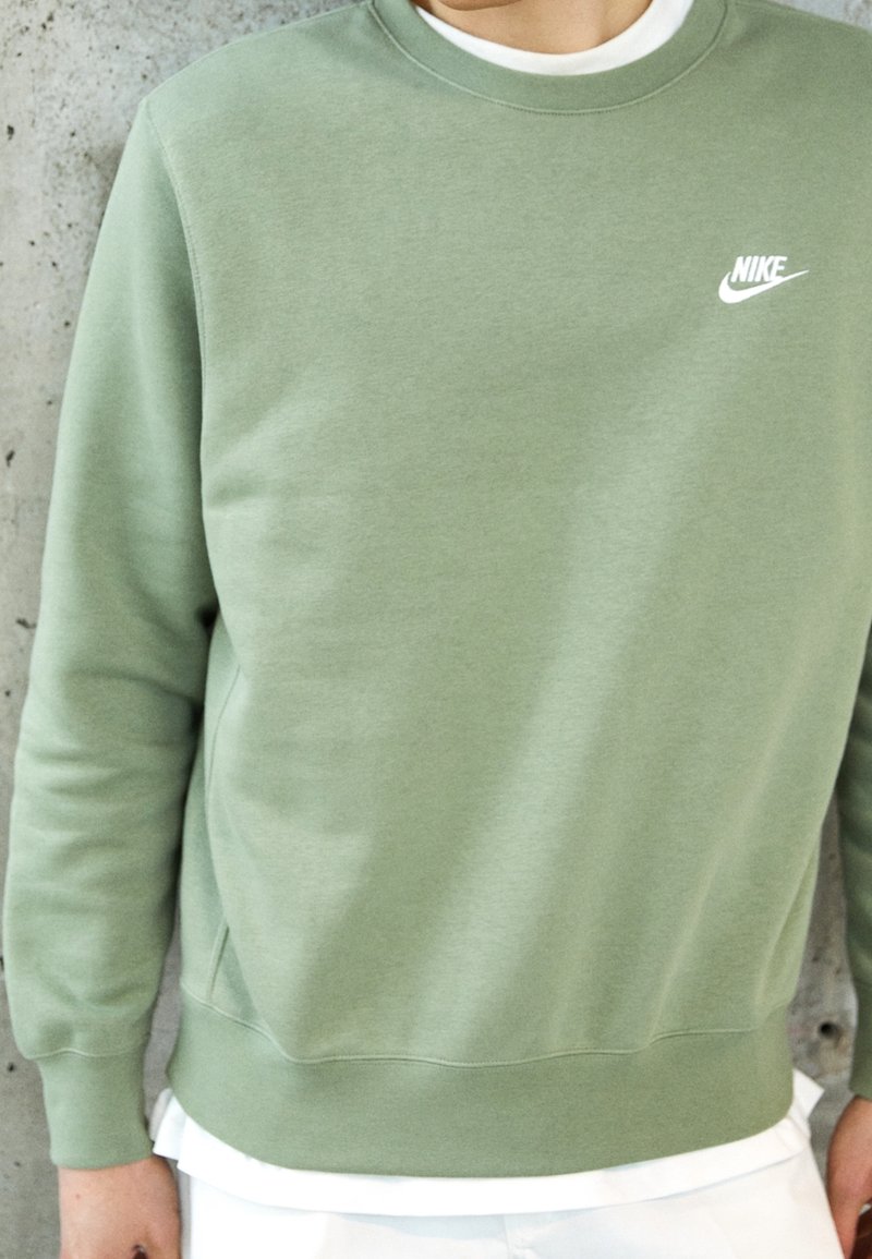 Personne portant un sweat-shirt Nike vert clair sur une chemise blanche, debout contre un mur en béton gris.