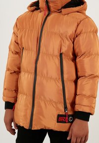 Chaqueta acolchada naranja con cremallera frontal, detalles en negro y una lengüeta roja. Incluye capucha y puños elásticos para mayor abrigo.