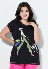 Camiseta negra con un gráfico de una mujer en un mono verde con estampado de leopardo, llevándolas bolsas de compras y paseando a un perro. Acentos en rosa.