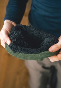 Contenedor redondo y suave en forro polar negro con un exterior de textura verde de punto. Sostenido en una mano, mostrando la profundidad y forma interior.