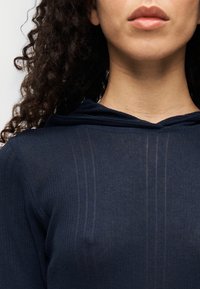 Μπλε ναυτικό φούτερ με κουκούλα που διαθέτει ριμπ ύφανση, εμφανή ραφές και άνετη εφαρμογή. Το ύφασμα φαίνεται ελαφρύ και ελαφρώς διαφανές.