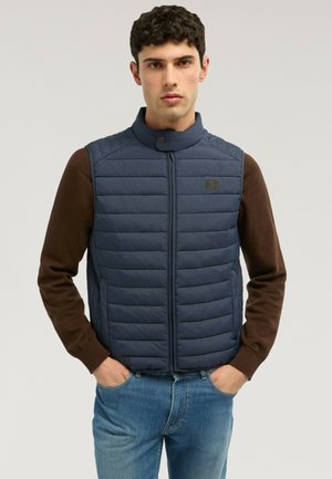 Młody mężczyzna w granatowej pikowanej kamizelce na brązowym swetrze i niebieskich dżinsach, stoi z rękami w kieszeniach na jednolitym tle.