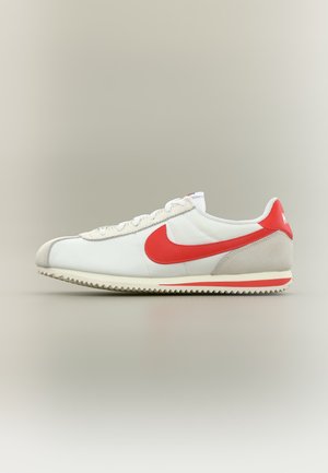 Hvid lavtop Nike sneaker med rød swoosh-logo, grå ruskindshæl-accenter og cremefarvet ydersål med rød stribe på en neutral baggrund.