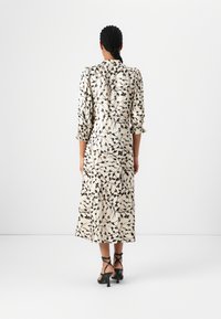 Langes, cremefarbenes Kleid mit schwarzem botanischem Druck, strukturierter Stoff, Puffärmel und einem fließenden Saum. Dazu stylische schwarze Sandalen mit Riemchen.