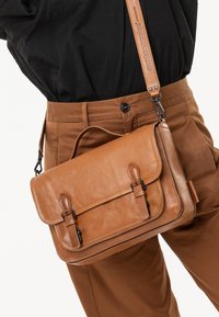 Sacoche en cuir marron avec des sangles à boucle et une bandoulière ajustable portée en travers sur un pantalon marron et un haut noir.