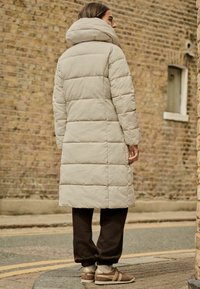 Lång beige vadderad kappa med hög krage, matchad med mörkbruna sweatpants och bruna sneakers. Bakgrund: tegelvägg och beläggning.