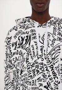 Personne portant un sweat à capuche blanc avec un texte noir de style manuscrit partout, incluant des mots comme "Versace Jeans Couture" et "sac."