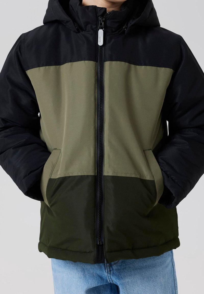 Personne portant une doudoune color-block noire, vert olive et vert foncé, les mains dans les poches, et un jean bleu clair.