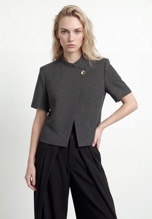Une femme blonde porte une veste gris anthracite à manches courtes, ornée d’un bouton doré, et un pantalon noir ample, sur un fond blanc uni.