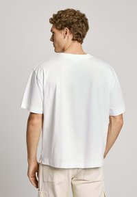 Witte katoenen t-shirt met een ontspannen pasvorm, korte mouwen, ronde hals en zijsplitten, weergegeven vanuit de achterkant. Zachte textuur.