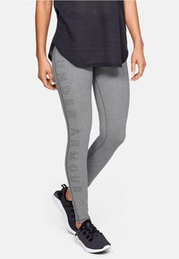 Szare legginsy z pionowym nadrukiem "Under Armour" w czerni, połączone z luźną czarną bluzą i czarnymi butami sportowymi. Gładki materiał i dopasowany fason.