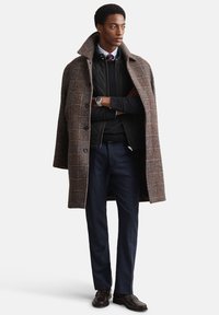 Bruine geruite overjas met brede kraag, gedragen over een zwart doorgestikt vest en marineblauwe broek; voorzien van een zilveren horloge en donkere schoenen.