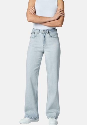 Person iført lyseblå jeans med høj talje og lige ben, hvid ærmeløs top og hvide sneakers, stående med armene krydset foran en ensfarvet baggrund.