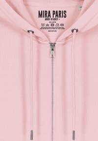 Sudadera con cremallera de un suave color rosa claro, hecha de 100% algodón. Presenta una cremallera plateada, cordones y una etiqueta con instrucciones de cuidado y el nombre de la marca.
