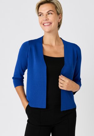 Mujer con cabello rubio corto sonriente, vestida con un cárdigan azul real abierto al frente sobre una camiseta negra y pantalones negros.