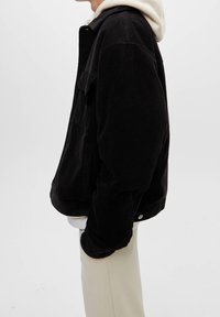 Giacca in velluto a coste nera con una vestibilità rilassata, dotata di una tasca frontale e accessori in metallo argentato. Indossata sopra un felpa grigia chiara e pantaloni beige.