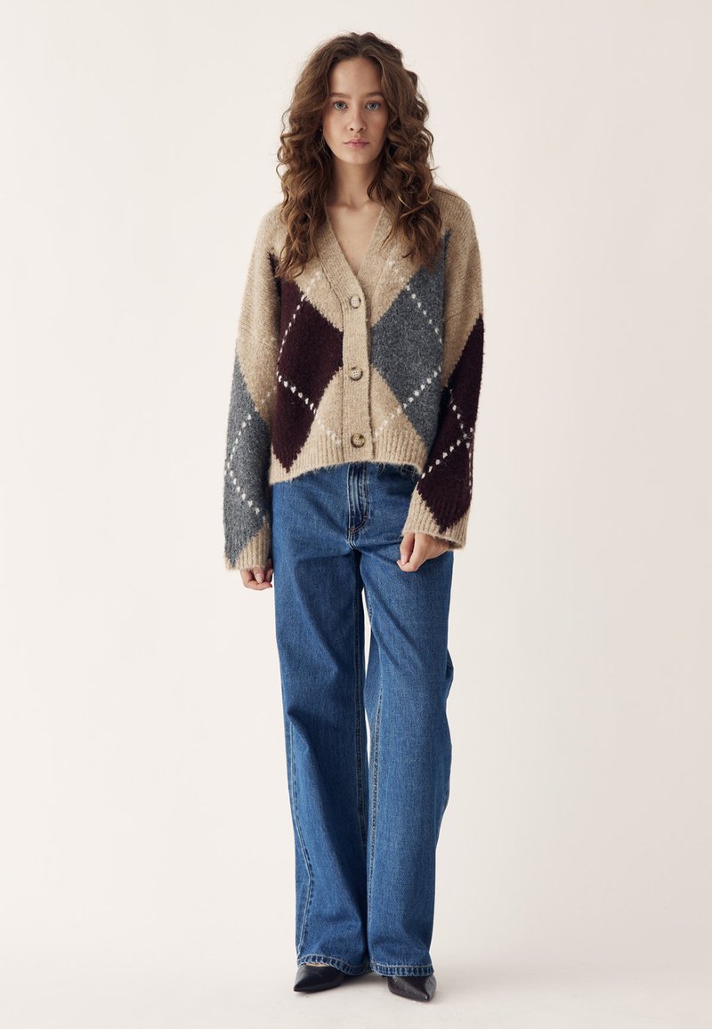 Noor naine lokkis juustega, kandes beeži, halli ja burgundia argyle-mustriga kardigani, siniseid laiade säärega teksapükse ja musti kingi seisab tavalise tausta ees.