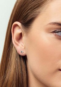 Rund, silverfärgad studsörhänge med en klar, facetterad sten, inställd i en enkel klordesign, buren på en modells öra.