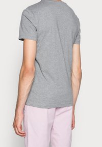 Camiseta gris de manga corta, cuello redondo, hecha de material de algodón suave. El fondo es liso, y la persona lleva puestos pantalones cortos de color rosa claro.