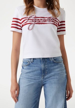 Kobieta nosząca białą krótka koszulkę z czerwonymi poziomymi paskami i napisami przypominającymi sznurek, w połączeniu z niebieskimi jeansami z wysokim stanem.