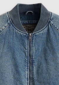 Chaqueta bomber de denim, azul, con cuello y puños acanalados, cierre de cremallera y etiqueta interior con detalles de marca. La textura parece suave con acentos desgastados.