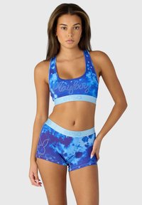 Modrá tie-dye športová podprsenka s textom "Playboy" a striebornými akcentmi; zodpovedajúce šortky majú logo a podobný vzor. Mäkká, elastická látka.