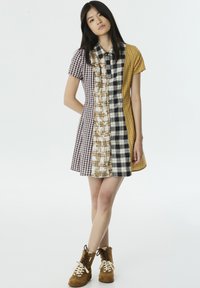 Vestido de manga corta con un diseño de patchwork que presenta varios patrones de cuadros en tonos de púrpura, amarillo y blanco. Llevado con botas beige con cordones.