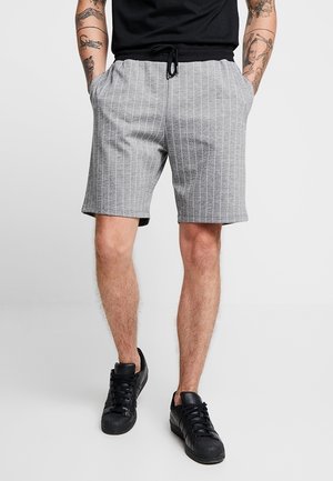 Hombre con pantalones cortos grises con rayas finas y cordón, zapatillas negras y camisa negra con los brazos tatuados visibles.