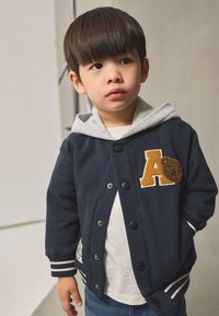 Giacca blu navy con cappuccio grigio, con strisce bianche sui polsini, una toppa a forma di "A" e un dettaglio a forma di biscotto textured. Indossata sopra una camicia bianca.