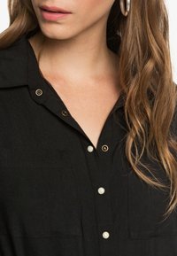 Camisa negra de botones con cuello, botones de perla y tono dorado, y una tela texturizada con dos bolsillos frontales.