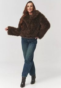Brązowa kurtka z imitacji futra, krótka, z dużym kołnierzem i szerokimi rękawami, połączona z niebieskimi, prostymi dżinsami i czarnymi botkami za kostkę.