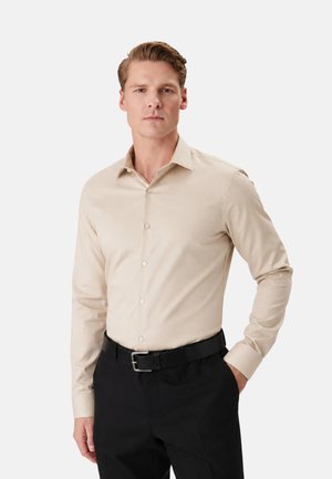 Hombre con camisa beige de manga larga abotonada metida en pantalones negros con cinturón negro, de pie con una mano en el bolsillo.