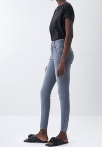 Vaqueros grises de corte ajustado con cintura alta, cierre de botones y un sutil desgaste. Combinados con una camiseta negra de manga corta y sandalias negras tipo slide.