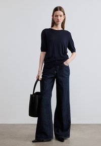 Ναυτικό μπλε πλεκτό τοπ με κοντά μανίκια, φαρδιά σκουρόχρωμα τζιν παντελόνια, μαύρη δερμάτινη τσάντα και παπούτσια μυτερά σε ουδέτερο φόντο.