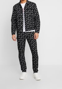 Svart jacka och byxor set med ett upprepat vitt logomönster, med en jacka med krage och figursydd stil. Vita sneakers kompletterar looken.