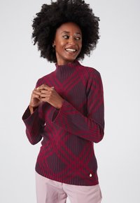 Maglione attillato color bordeaux con texture a coste e un motivo a quadri rossi a contrasto. Presenta maniche a campana e un piccolo logo in accentuazione.