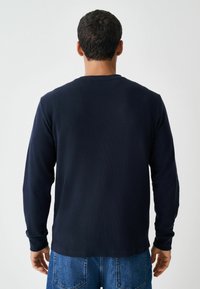Marineblauwe longsleeve shirt met een gestructureerde stof en een ronde halslijn, gecombineerd met lichtblauwe denim jeans. Zichtbare mouwoevers en ontwerpdetails aan de achterkant.