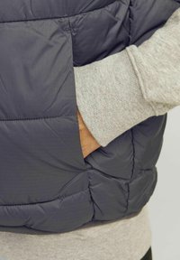 Donkergrijze puffervest met een gestikt patroon en zijzakken, gelaagd over een lichtgrijze sweatshirt met geribde manchetten.