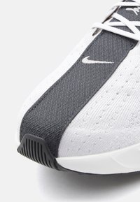 Primer plano de la puntera de una zapatilla Nike blanca y gris oscuro con material de punto texturizado, cordones blancos y una suela de goma negra.