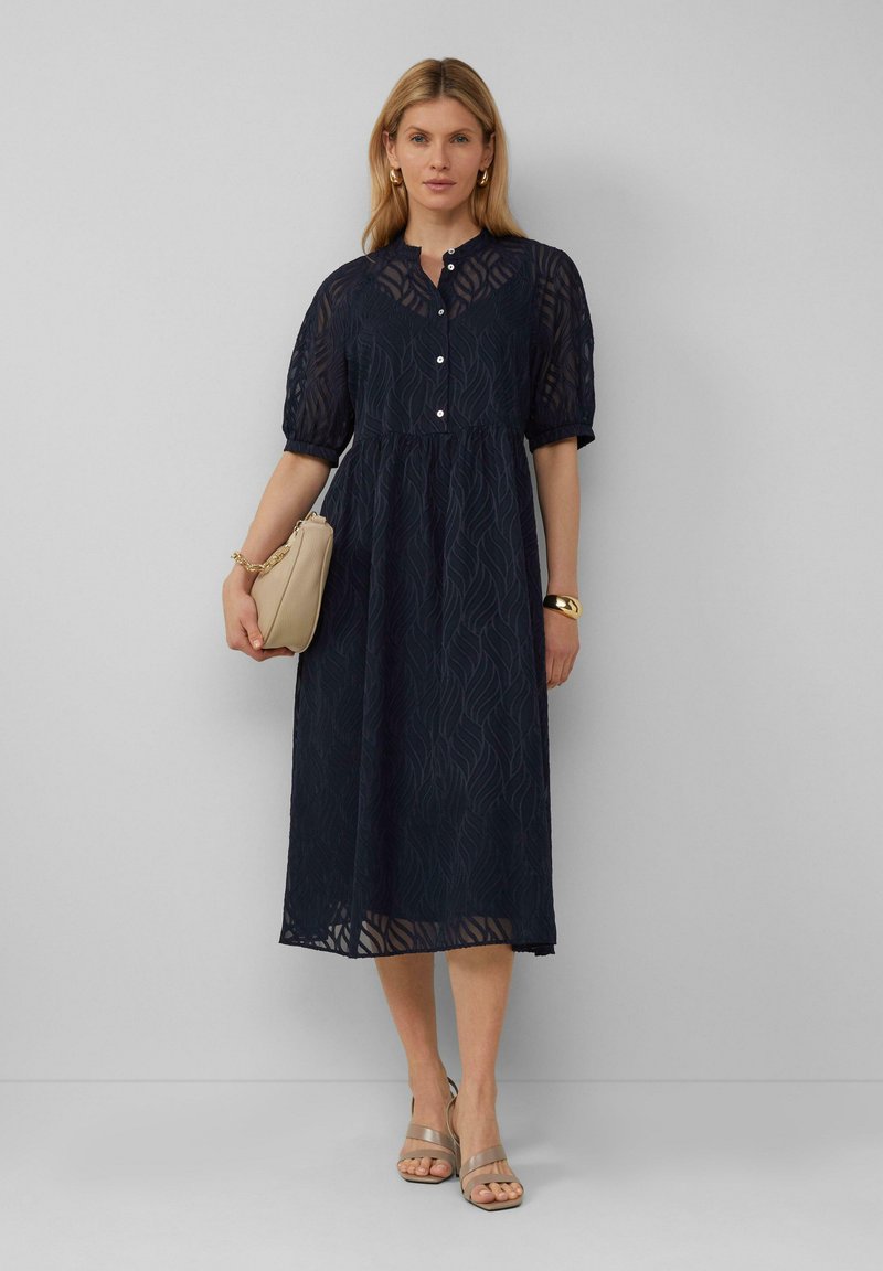 Robe midi bleu marine avec un motif de vagues texturé, manches bouffantes courtes, décolleté à boutons et silhouette évasée. Sac à main beige et sandales.
