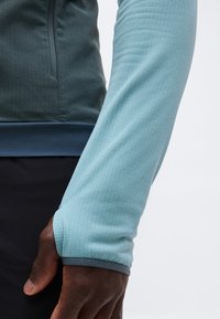 Κοντινό πλάνο σκοτεινής επιδερμίδας χεριού που φοράει ανοιχτό γαλάζιο μπλουζάκι μακρυμάνικο με άνοιγμα στον αντίχειρα, σκούρο παντελόνι και σκούρο μπουφάν με φερμουάρ.