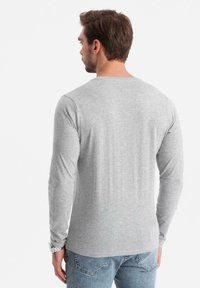 Man med ljusgrå långärmad skjorta och blå jeans, sedd bakifrån mot en enfärgad vit bakgrund.