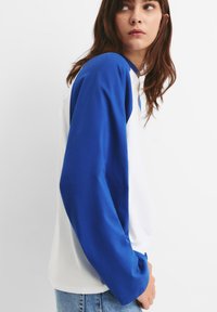 Bela in modra majica z dolgimi rokavi, ki ima raglan rokave in mehak material. Telo je belo, rokavi in ovratnik pa sta svetlo modra.
