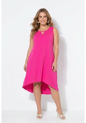 Donna che indossa un abito smanicato rosa acceso con balze asimmetriche e sandali bianchi con tacco, in piedi davanti a uno sfondo bianco uniforme.